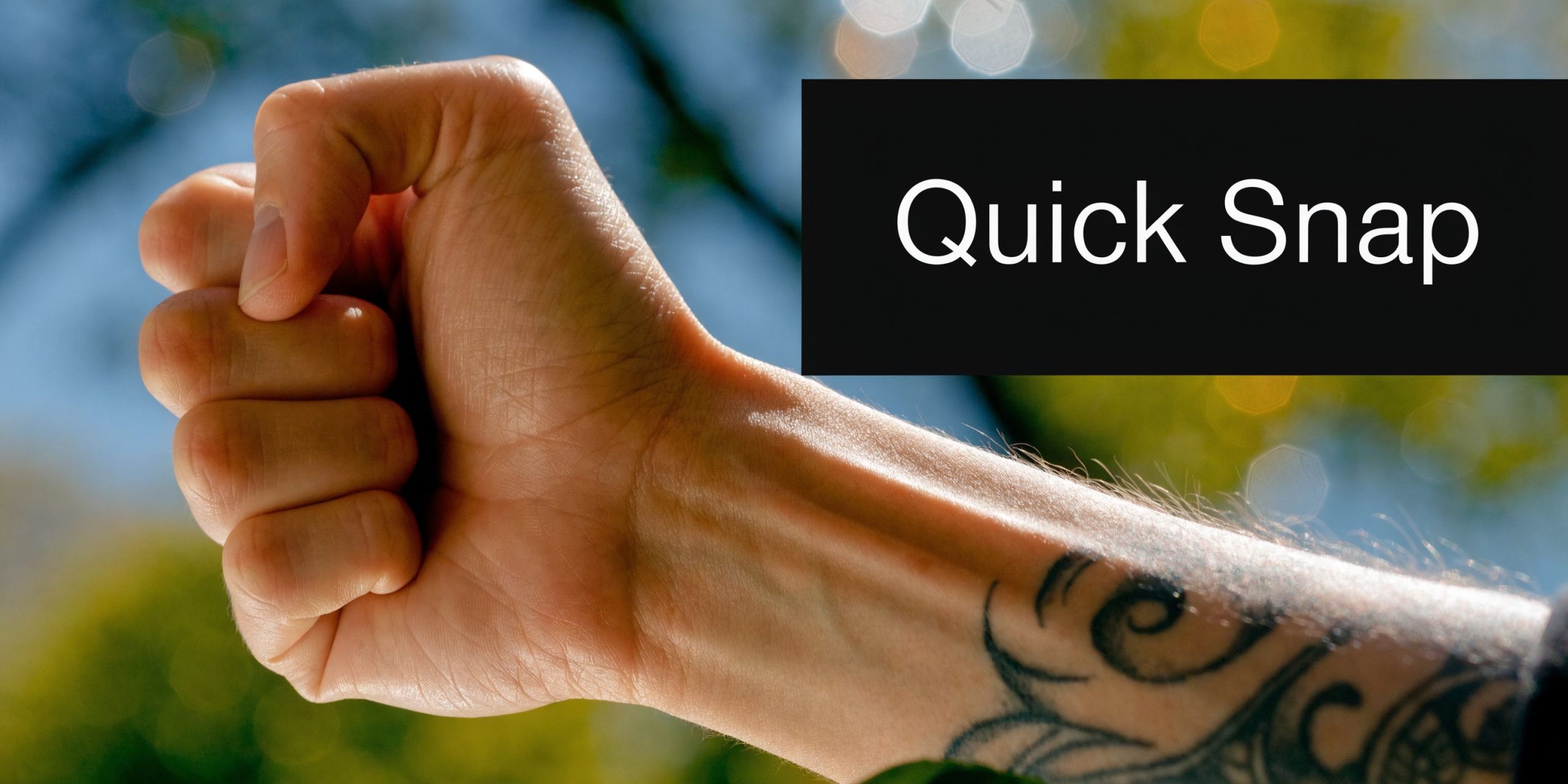 A close-up view of a clenched fist with a visible forearm tattoo against a blurred outdoor background.