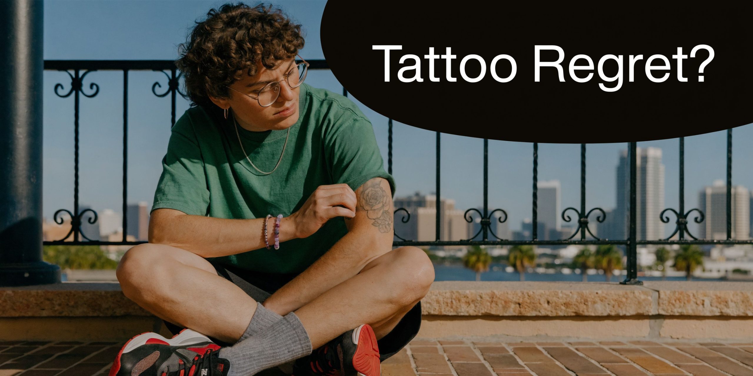 A person with curly hair sitting on a rooftop overlooking a city while inspecting their arm tattoo.