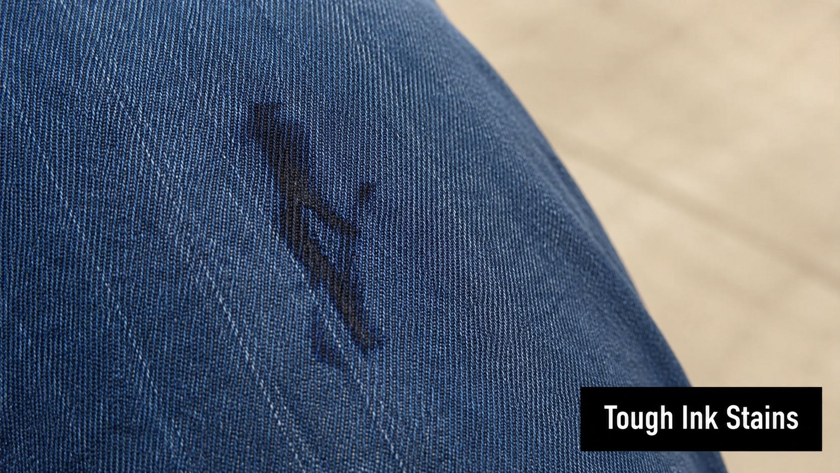 A close-up view of a dark ink stain on blue denim fabric, clearly showing the fabric texture.